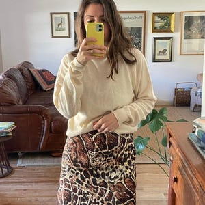 May include: A long-sleeved cream-colored sweater with a crew neckline paired with a short skirt featuring a brown, black, and tan animal print pattern. The outfit is worn over solid black leggings, creating a layered look with neutral tones.