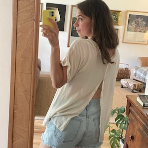 Könnte beinhalten: Ein helles Oberteil mit offenem Rücken und hellblaue Jeansshorts. Das Oberteil hat kurze Ärmel und eine lockere Passform. Die Shorts sind im Used-Look mit ausgefransten Kanten. Das Outfit spiegelt sich in einem Holzrahmen wider.