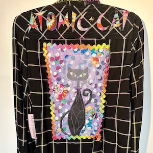 May include: Black and white checked flannel shirt with a colourful patch on the back. The patch features a black cat with a purple background and a rainbow border. The text "ATOMIC CAT" is embroidered on the back of the shirt.