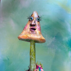 May include: A whimsical mushroom figurine with a painted face, green eyes, and a pink mouth. The mushroom is in a brown pot with a pink and green flower on top. The figurine is against a green and blue background.