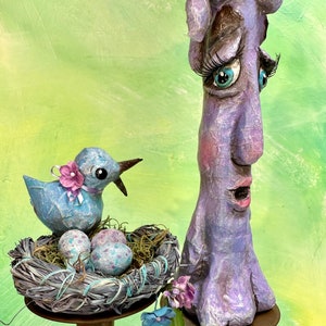 May include: A blue bird with a pink bow sits in a nest with two blue and white speckled eggs. The bird is perched on a gold candlestick. A purple, textured, human-like figure with blue eyes and eyelashes stands on a gold candlestick.