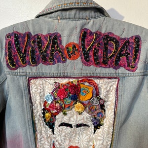May include: A light blue denim jacket with a colorful embroidered patch featuring a portrait of Frida Kahlo. The patch is surrounded by a yellow border and the words "Viva la Vida!" are embroidered in black and pink on the top of the jacket.
