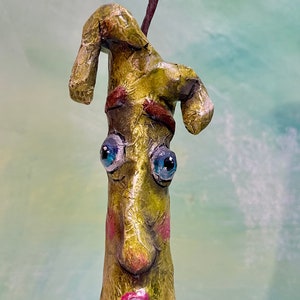 May include: A whimsical candle holder with a green, textured, human-like face, blue eyes, and a pink mouth. The candle holder is topped with a small, yellow, glittery flame and a blue and white flower.