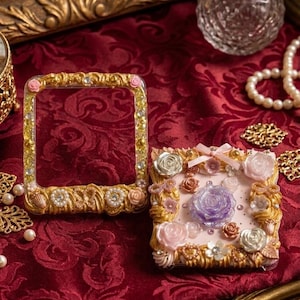 May include: Two ornate, gold-colored picture frames with floral and jewel embellishments sit on a red patterned surface. One frame is square, the other rectangular. A small perfume bottle and pearls are also visible.