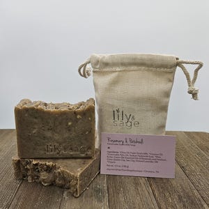 Può includere: Due saponette di latte di capra marrone impilate l'una sull'altra. Il sapone è etichettato "lilysage". Il sapone è accanto a una piccola borsa di mussola bianca e un biglietto da visita rosa che recita "Rosemary & Patchouli Handmade Goat's Milk Soap" e elenca gli ingredienti.