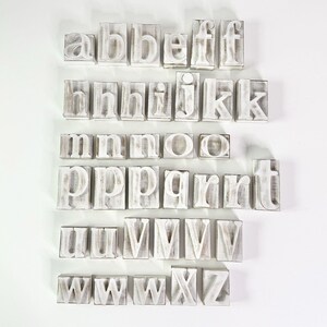 May include: A collection of distressed, white-painted wooden alphabet letters. Each letter is individually mounted on a rectangular block. The letters are arranged in rows, showcasing the full alphabet from A to Z.