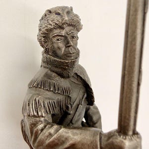 May include: A metallic silver-toned figurine depicting a figure wearing a fur hat with an animal head design and a fringed jacket. The figure has a high collar and is holding a long, slender staff or pole in a vertical position, rendered in a monochromatic finish.