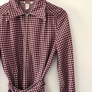 Puede incluir: Vestido a cuadros burdeos y blancos con cuello y mangas largas. El vestido tiene una cremallera de largo completo y un cintur&oacute;n de tela a juego que se ata a la cintura. El vestido est&aacute; colgado de una percha de madera.