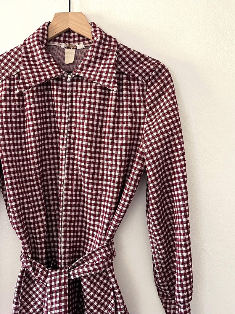 Puede incluir: Vestido a cuadros burdeos y blancos con cuello y mangas largas. El vestido tiene una cremallera de largo completo y un cintur&oacute;n de tela a juego que se ata a la cintura. El vestido est&aacute; colgado de una percha de madera.