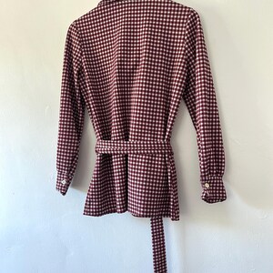 Puede incluir: Chaqueta a cuadros burdeos y blancos con mangas largas y cintur&oacute;n a juego. La chaqueta tiene cuello camisero y pu&ntilde;os abotonados. La prenda est&aacute; colgada de una percha negra contra una pared blanca.