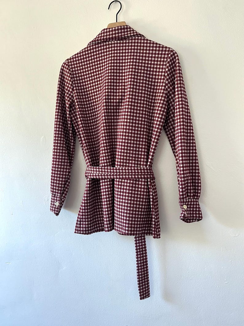 Puede incluir: Chaqueta a cuadros burdeos y blancos con mangas largas y cintur&oacute;n a juego. La chaqueta tiene cuello camisero y pu&ntilde;os abotonados. La prenda est&aacute; colgada de una percha negra contra una pared blanca.