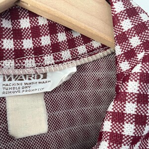 Puede incluir: Primer plano de una tela con estampado de cuadros burdeos y blancos, posiblemente una prenda vintage. La tela est&aacute; drapeada sobre una percha de madera. Una etiqueta con instrucciones de cuidado es visible, incluyendo "MACHINE WASH WARM" y "TUMBLE DRY".