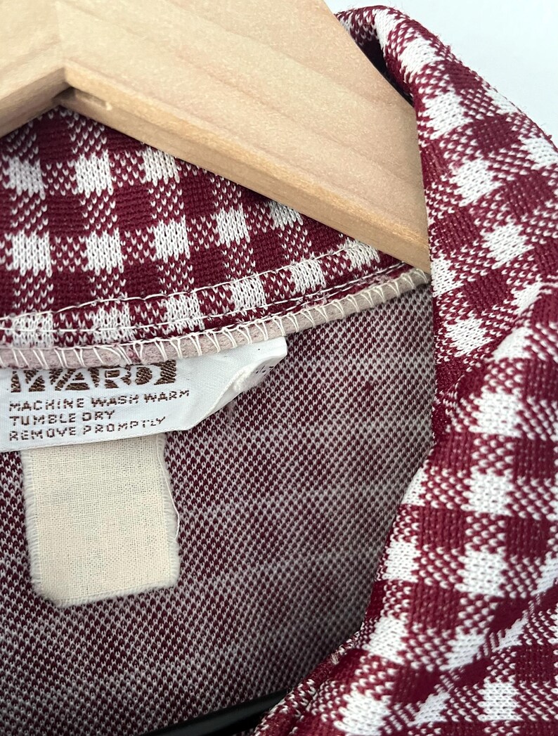 Puede incluir: Primer plano de una tela con estampado de cuadros burdeos y blancos, posiblemente una prenda vintage. La tela est&aacute; drapeada sobre una percha de madera. Una etiqueta con instrucciones de cuidado es visible, incluyendo "MACHINE WASH WARM" y "TUMBLE DRY".