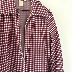 Puede incluir: Chaqueta con cremallera a cuadros burdeos y blancos colgada de una percha de madera. La chaqueta tiene un cuello ancho y mangas largas. La cremallera es plateada. La tela tiene un estampado de cuadros vichy.