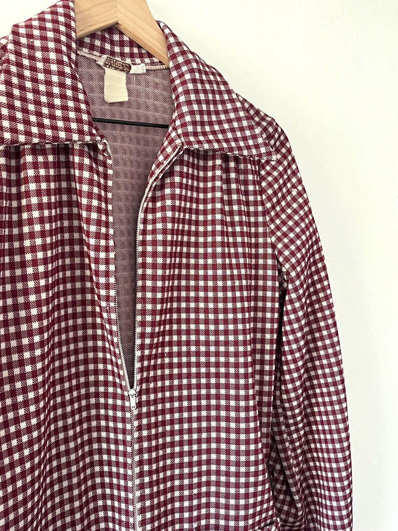 Puede incluir: Chaqueta con cremallera a cuadros burdeos y blancos colgada de una percha de madera. La chaqueta tiene un cuello ancho y mangas largas. La cremallera es plateada. La tela tiene un estampado de cuadros vichy.