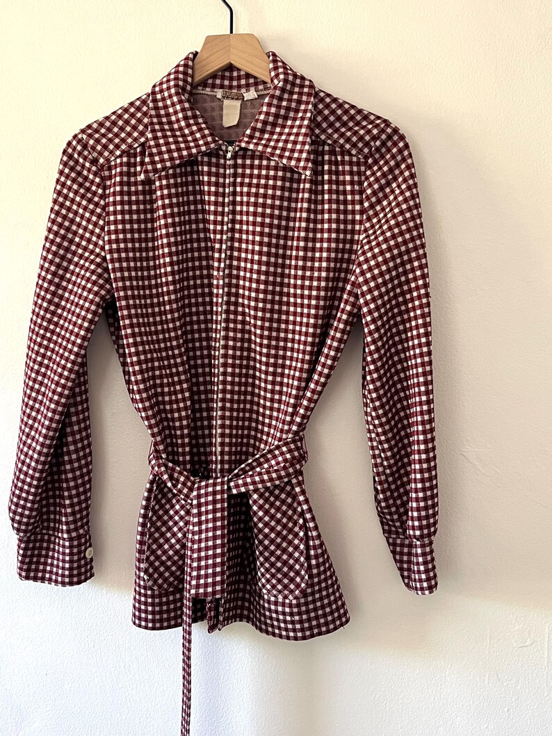 Puede incluir: Top a cuadros de color burdeos y blanco, con mangas largas y cintur&oacute;n anudado. El top tiene un cuello con solapas y una cremallera de largo completo. La prenda est&aacute; colgada de una percha de madera contra una pared blanca.