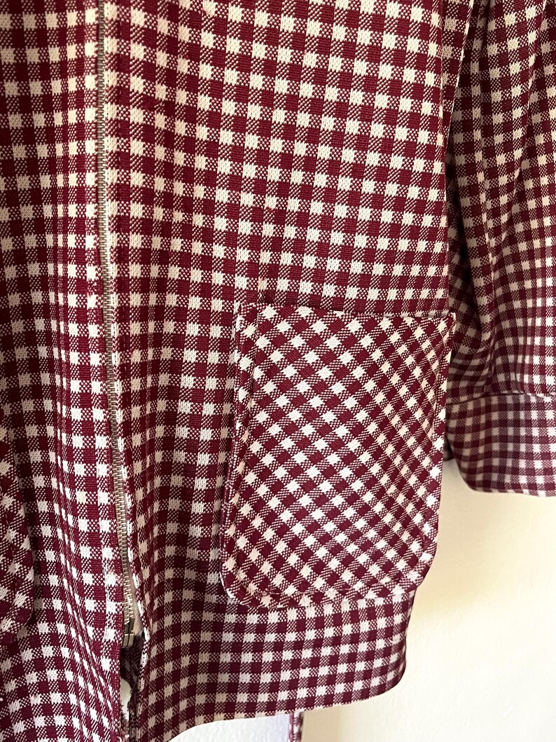 Puede incluir: Primer plano de una chaqueta a cuadros burdeos y blancos con cremallera plateada y bolsillo. El patr&oacute;n de la chaqueta es un dise&ntilde;o cl&aacute;sico de cuadros vichy, con el bolsillo que refleja el mismo patr&oacute;n. El pu&ntilde;o de la manga es visible.