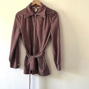 Puede incluir: Una camisa de manga larga a cuadros burdeos y blancos con cintur&oacute;n a juego. La camisa est&aacute; colgada en una percha de madera contra una pared blanca. La camisa tiene cuello y cremallera.