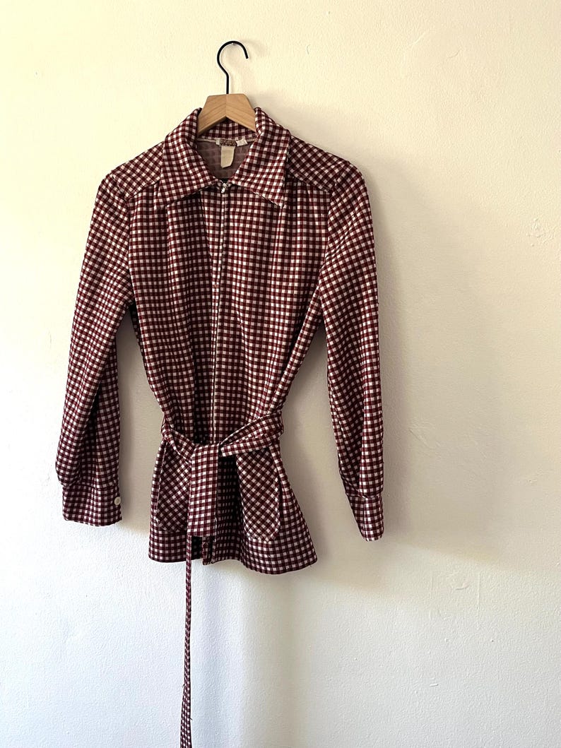 Puede incluir: Una camisa de manga larga a cuadros burdeos y blancos con cintur&oacute;n a juego. La camisa est&aacute; colgada en una percha de madera contra una pared blanca. La camisa tiene cuello y cremallera.