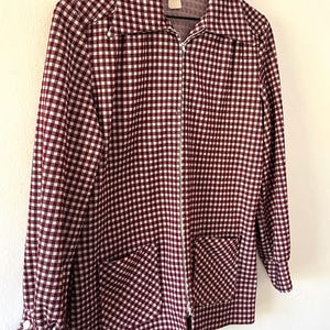 Puede incluir: Chaqueta con cremallera a cuadros burdeos y blancos, con dos bolsillos delanteros y mangas largas. La chaqueta est&aacute; colgada de una percha de madera sobre un fondo blanco. El cuello est&aacute; abierto y la cremallera es plateada.