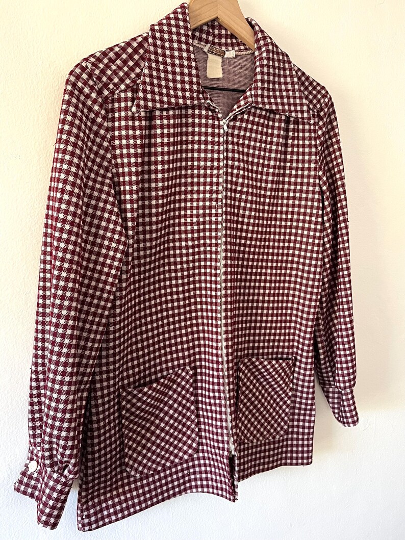 Puede incluir: Chaqueta con cremallera a cuadros burdeos y blancos, con dos bolsillos delanteros y mangas largas. La chaqueta est&aacute; colgada de una percha de madera sobre un fondo blanco. El cuello est&aacute; abierto y la cremallera es plateada.