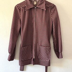 Puede incluir: Chaqueta a cuadros burdeos y blancos con cremallera completa, dos bolsillos delanteros y cintur&oacute;n a juego. La chaqueta est&aacute; colgada de una percha de madera contra una pared blanca. El patr&oacute;n de la chaqueta es un dise&ntilde;o cl&aacute;sico de cuadros vichy.