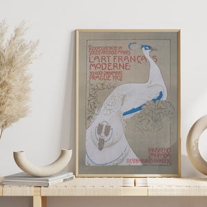Op de afbeelding: Een ingelijste vintage art nouveau poster met een witte pauw met blauwe accenten. De poster, met Franse tekst, adverteert een kunsttentoonstelling in Praag in 1902. Het wordt getoond op een houten bank met decoratieve vazen en gedroogde planten.