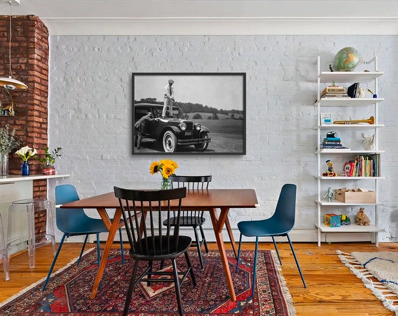 Op de afbeelding: Een zwart-witfoto van een man die op de treeplank van een vintage auto staat. De auto staat geparkeerd op een grasveld. De man draagt een wit overhemd en een witte broek en een hoed. De auto heeft een reservewiel achterop en een kentekenplaat met de tekst "3317".