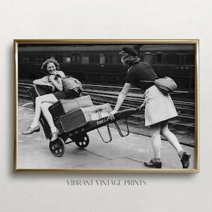 May include: Two women in vintage clothing stand on a platform with luggage. One woman is pushing a luggage cart with suitcases on it. The cart has a sign that says "Tare 1-0-7".