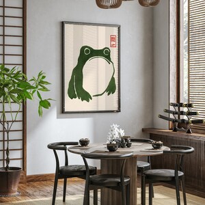 May include: A dining room with a round wooden table and six black chairs. A framed print of a green frog with a red stamp on the wall. A potted plant in the corner.