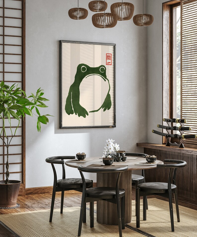 May include: A dining room with a round wooden table and six black chairs. A framed print of a green frog with a red stamp on the wall. A potted plant in the corner.