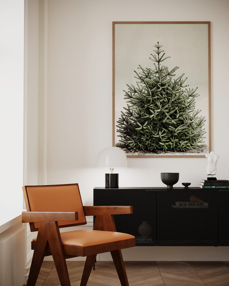 May include: A framed botanical print of a green evergreen tree hangs above a black cabinet. A mid-century modern chair with a brown wooden frame and orange leather seat sits in front of the cabinet. A white lamp with a black base is on the cabinet.