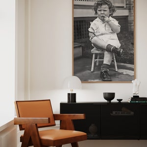 Peut inclure: Une photographie en noir et blanc d'un jeune enfant assis sur un petit tabouret en bois. L'enfant porte une chemise &agrave; manches courtes et un short. L'enfant tient un petit objet dans sa main et regarde la cam&eacute;ra.