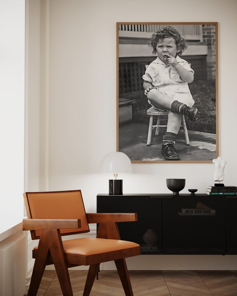 Peut inclure: Une photographie en noir et blanc d'un jeune enfant assis sur un petit tabouret en bois. L'enfant porte une chemise &agrave; manches courtes et un short. L'enfant tient un petit objet dans sa main et regarde la cam&eacute;ra.