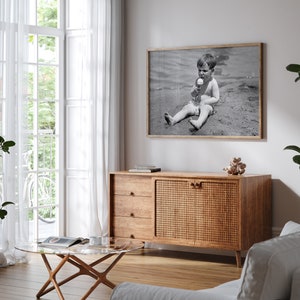 May include: A wooden cabinet with a woven rattan door and three drawers. The cabinet is in a living room with a white couch, a glass coffee table, and a black and white photo of a child on the beach.