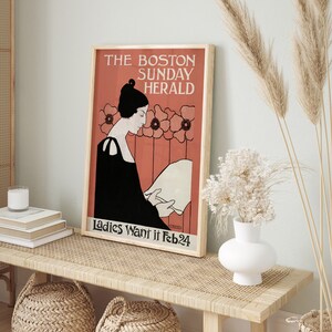 May include: Framed vintage poster with the text "THE BOSTON SUNDAY HERALD" and "Ladies Want it Feb24". The poster features an illustration of a woman in a black dress reading a paper, set against a coral background with floral accents. The poster is displayed on a light wood bench.