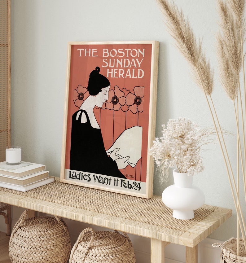 May include: Framed vintage poster with the text "THE BOSTON SUNDAY HERALD" and "Ladies Want it Feb24". The poster features an illustration of a woman in a black dress reading a paper, set against a coral background with floral accents. The poster is displayed on a light wood bench.