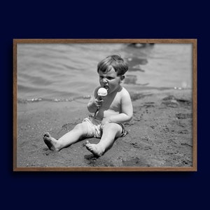 May include: A young boy sits on a sandy beach, wearing a pair of shorts and eating an ice cream cone.