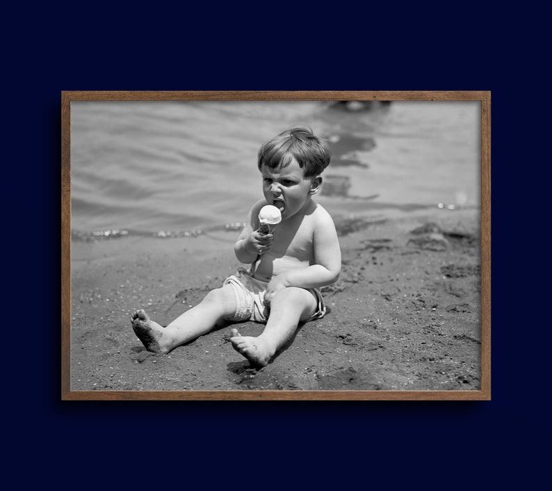May include: A young boy sits on a sandy beach, wearing a pair of shorts and eating an ice cream cone.