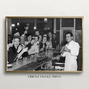 May include: A black and white photograph of a group of people at a bar. The people are all smiling and raising their glasses in a toast. The bartender is shaking a cocktail shaker. The image is framed in a gold frame. The text 