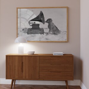 May include: A black and white photograph of a dog listening to a vintage gramophone. The dog is sitting in the snow and the gramophone is on a wooden box. The photograph is framed in a light wood frame and hanging on a light beige wall above a wooden cabinet.