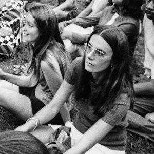 Puede incluir: Dos mujeres sentadas al aire libre, una con el pelo largo y oscuro que lleva gafas redondas y una camiseta gris, la otra con el pelo largo y claro que lleva una camiseta oscura.