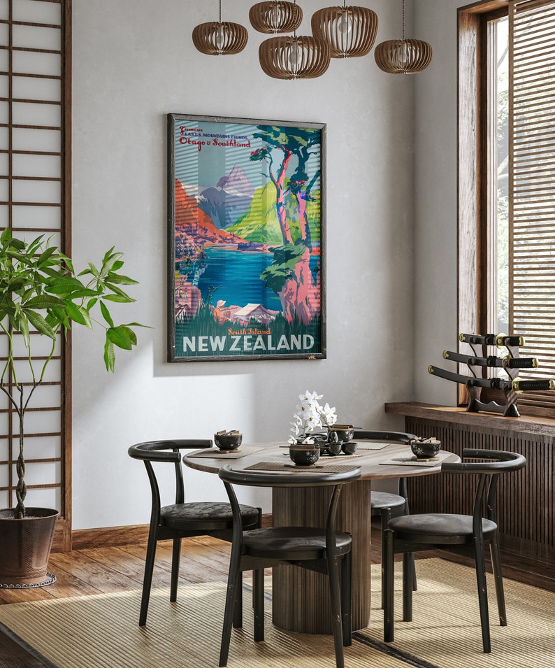 May include: A vintage travel poster for New Zealand featuring a scenic landscape with mountains, a lake, and trees. The poster is framed and hanging on a wall in a dining room with a wooden table and black chairs.
