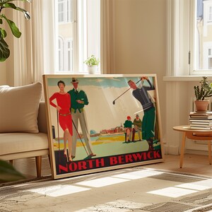 May include: A framed vintage-style poster with the text "NORTH BERWICK" in red. The poster depicts a golf scene with figures in period attire, including a woman in a red dress and a man in a green jacket. The frame is a light wood color.