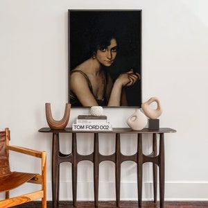 Pode incluir: Uma mesa de consola de madeira escura com uma pintura emoldurada em preto de uma mulher com cabelo escuro e um brinco de p&eacute;rola branca. A pintura est&aacute; sobre uma pilha de livros com o t&iacute;tulo "TOM FORD 002" vis&iacute;vel. H&aacute; duas esculturas de cer&acirc;mica na mesa, uma em forma de vaso e a outra em forma de U.
