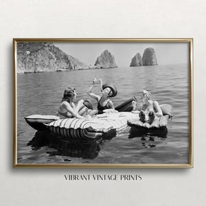 May include: A black and white photograph in a gold frame depicting three individuals on inflatable rafts in the ocean. They are gathered around a floating table with drinks and food, with large rock formations visible in the background.