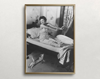 Puede incluir: Fotografía enmarcada en blanco y negro de una mujer estirándose en la cama. Lleva un conjunto de dormir de color claro. Un gato salta en el suelo. La habitación tiene papel pintado floral y una cortina estampada.