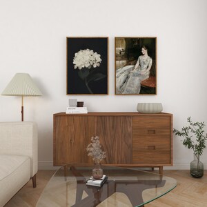 May include: Two framed art prints are displayed above a wooden cabinet. One shows a white hydrangea on a black backdrop. The other depicts a woman in a long dress. A lamp, sofa, and coffee table are in the foreground.