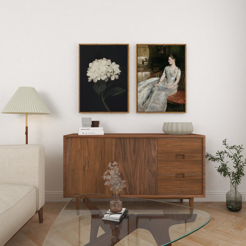 May include: Two framed art prints are displayed above a wooden cabinet. One shows a white hydrangea on a black backdrop. The other depicts a woman in a long dress. A lamp, sofa, and coffee table are in the foreground.