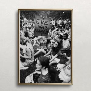May include: Black and white photograph of a group of people sitting on the grass. A sign reads "Eve Was Framed".